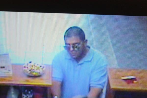 security camera image of man with blue polo shirt and sunglasses on, sitting at a table